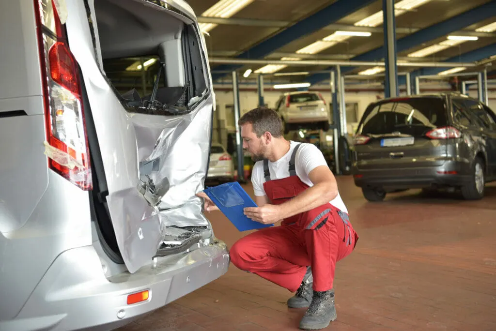 Ein Mann in roter Latzhose hockt in einer Werkstatthalle mit einem blauen Klemmbrett vor dem eingedrückten Heck eines silbernen Transporters, im Hintergrund stehen Autos auf Hebebühnen