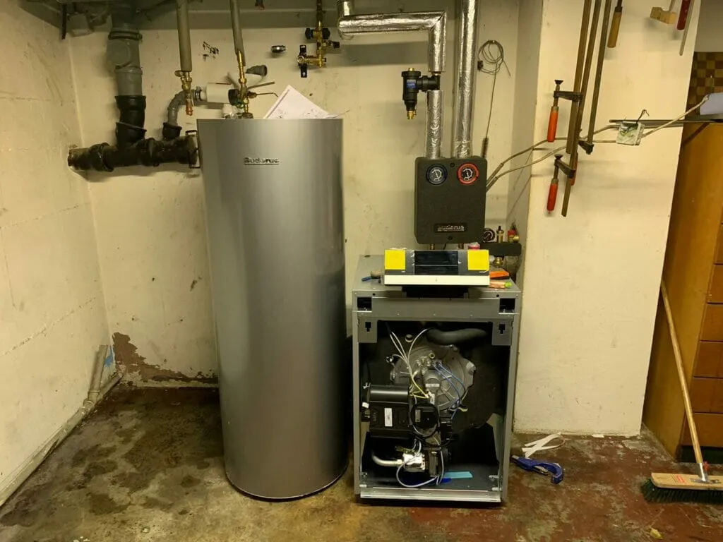 Heizkessel mit abgenommener Frontverkleidung neben einem silbernen Warmwasserspeicher in einem Kellerraum, darüber Rohrleitungen und eine Pumpengruppe