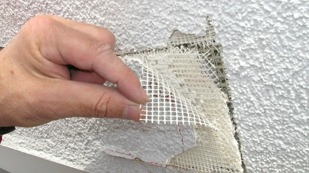 Hand zieht ein loses Armierungsgewebe von einer schadhaften Putzfassade ab