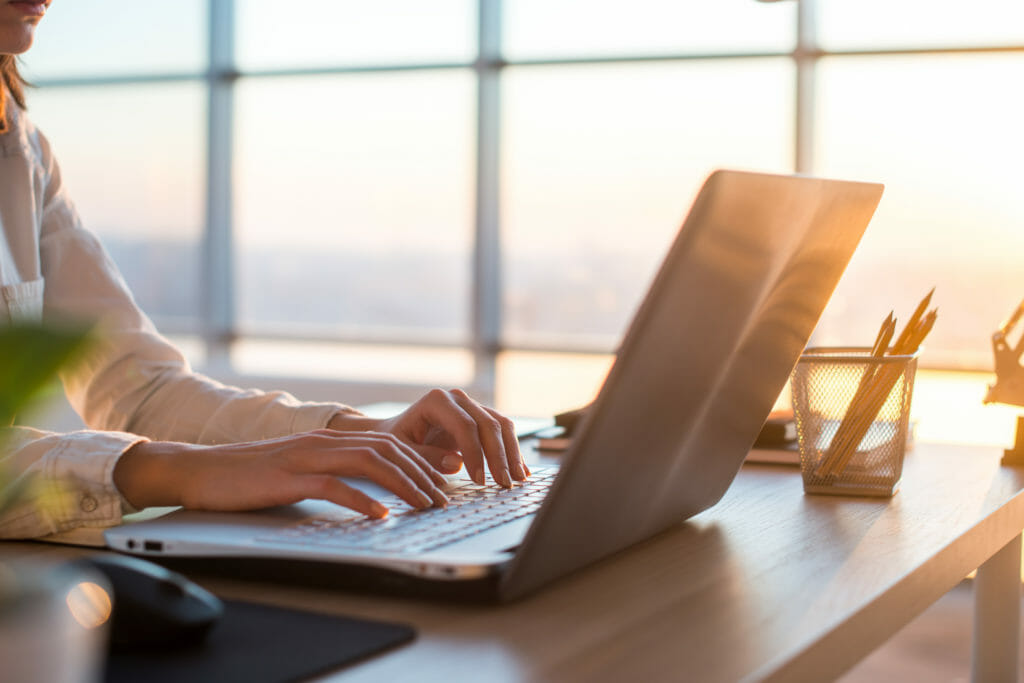 Frau arbeitet an einem Gutachten an einem Laptop am Schreibtisch mit Abendsonne im Hintergrund